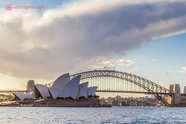 A Ópera de Sydney vista de um outro ângulo, do Jardim Botânico da cidade. O céu está parcialmente nublado, com uma enorme nuvem, mas com grandes partes azuis. A Sydney Bridge aparece atrás dela.