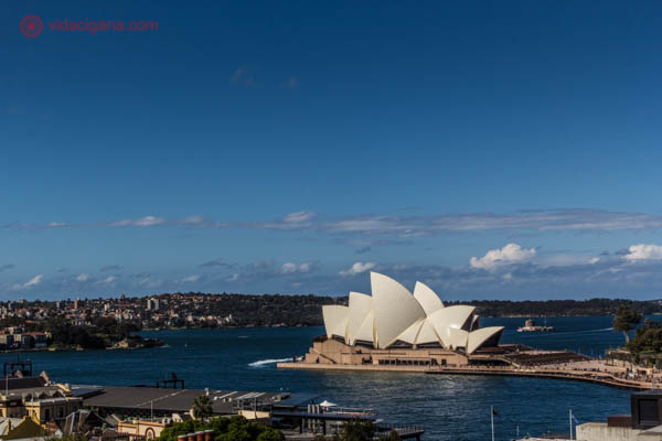 Onde ficar em Sydney: Vista do bairro The Rocks, em Sydney. Do alto de um prédio é possível ver a Ópera de Sydney bem em frente, com a baía azul. O céu está azul com algumas nuvens brancas.