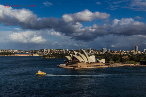 A Ópera de Sydney, com um barquinho amarelo passando em sua frente, com o céu azul e várias nuvens brancas.