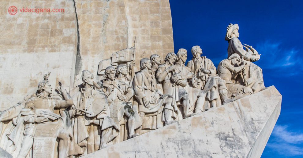 Detalhe do lado leste do Padrão dos Descobrimentos, em Lisboa, com vários navegadores e personalidades importantes para a expansão marítima portuguesa. O monumento é feito de pedra, e o céu ao fundo é azul.