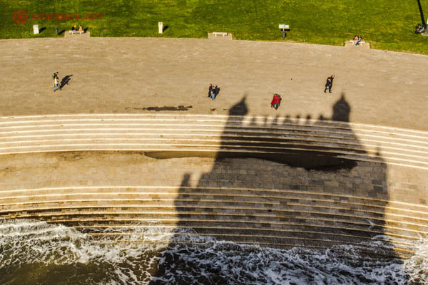 A sombra da Torre de Belém, em Lisboa, projetada em sua praia, com pessoas andando pelos degraus e as ondas batendo.