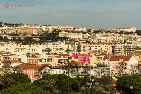O bairro de Belém, em Lisboa, com suas casas coloridas e telhados cor de ocre. As casas vão subindo pelas colinas. Na base da foto, várias ávores com folhas verdes se encontram. O céu está azul. Uma casa rosa se destaca.