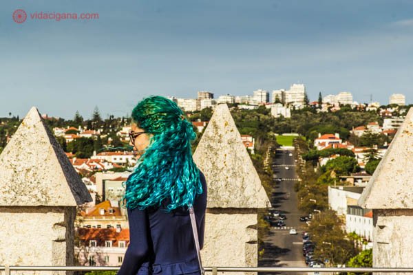 Uma menina com o cabelo em degradé do verde para o azul e encaracolado está no topo da Torre de Belém, em Lisboa. Ao fundo, a cidade, com ruas e casas e muito verde.