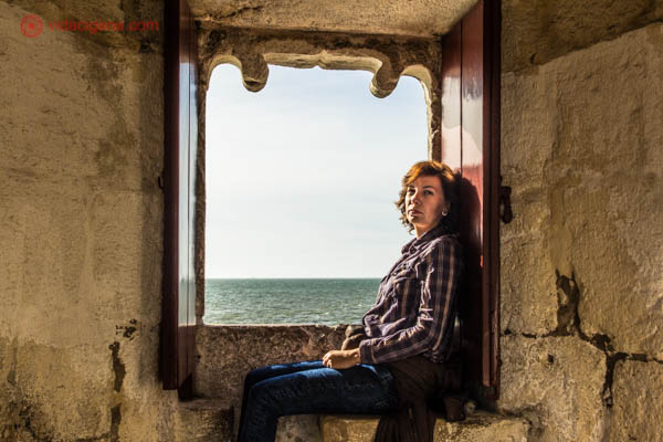 Uma mulher sentada na frente da janela da Torre de Belém em Lisboa, com o rio Tejo ao fundo. A foto é escura.