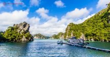 Halong Bay, no Vietnã, com suas lindas ilhas cheias de verde, suas águas mornas verdes, e seu céu azul. Com um barco ancorado e dois barquinhos amarrados em seu pier. Tudo o que você precisa saber sobre o clima no Vietnã e a melhor época para se visitar o país
