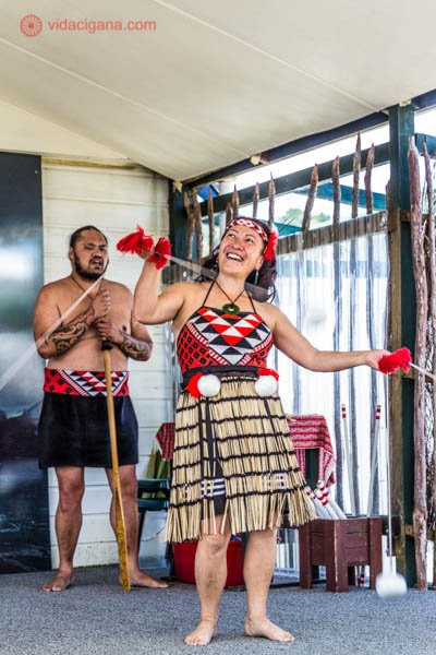 Uma mulher dançando a cultura maori na Nova Zelândia, com vestes típicas, vermelhas, pretas e com saia de folhas de árvores secas, chamada piupiu. Ela dança com bolinhas amarradas em barbantes.