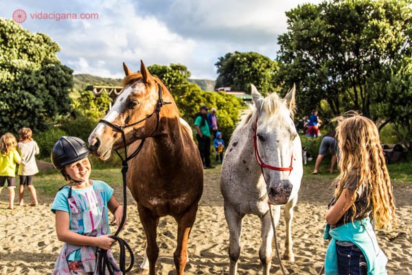 Duas meninas neozelandesas com dois cavalos, um marrom e outro branco. Elas usam capacetes para andar de cavalo e seguram os cavalos em cordas. O lugar tem areia e ao mesmo tempo muito verde em volta.