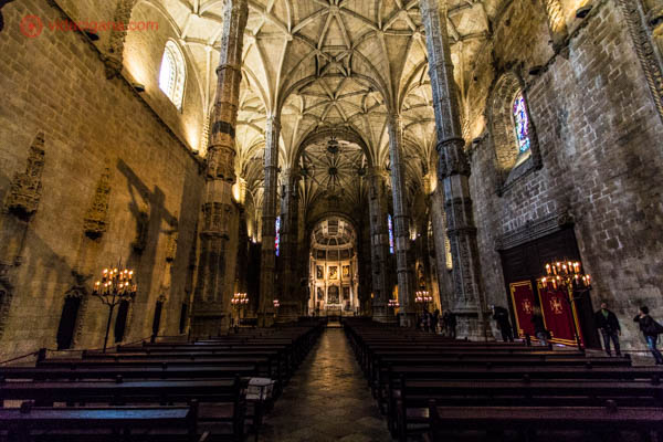 O interior da igreja do Mosteiro dos Jerônimos em Lisboa, com seu interior escuro e seu teto branco e abobadado. Muitos bancos de madeira vão até o altar. A luz entra por algumas janelas em seu topo.