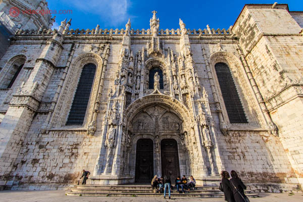 A fachada do Mosteiro dos Jerônimos em Lisboa com sua marcante arquitetura manuelina. Feito de pedras brancas, totalmente adornado. O céu está azul e algumas pessoas estão sentadas em seus degraus.