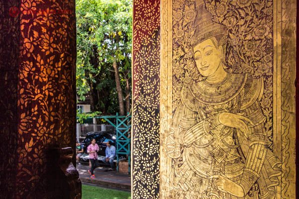 A porta de um templo em Chiang Mai, na Tailândia, em tons dourados e avermelhados. Uma imagem está esculpida na porta. Lá fora é possível ver entre as portas um casal conversando embaixo das árvores.