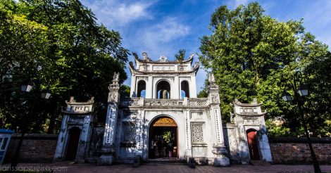 Um prédio histórico em Hanói na entrada do Templo da Literatura na capital do Vietnã. Ele possui arquitetura oriental, com as quinas curvadas para cima, é um prédio branco e pequeno que hoje mais serve como pórtico. A sua frente árvores com folhas verdes fazem sombra para quem passa. O céu está azul.