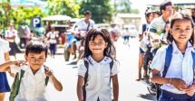 três crianças, duas meninas e um menino cambojanos andando pelas ruas de Phnom Penh, a capital do Camboja. eles voltam da escola pois estão os treês uniformizados. os três sorriem para a câmera.