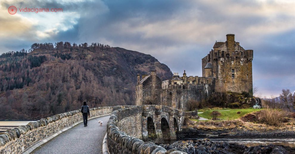 O castelo eilean donan, nos highlands da escócia, cercado por 3 lochs e por montanhas nevadas. Um homem caminha com um schnauzer branco em direção ao castelo. Está anoitecendo e um pequeno por do sol pode ser visto em meio as nuvens.