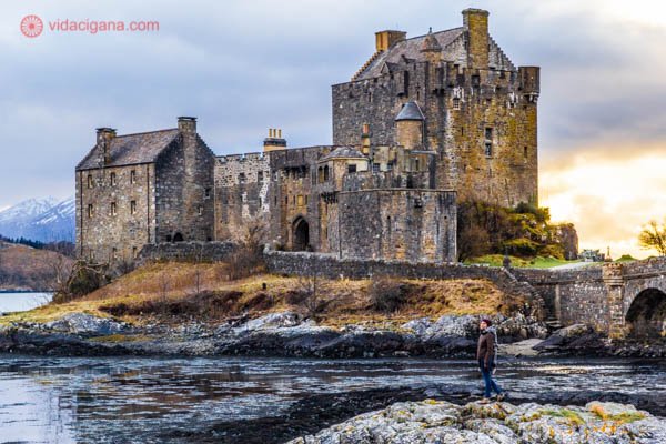 O Castelo Eilean Donan em zoom, com uma mulher de pé em frente a ele. O dia está nublado e bem frio.