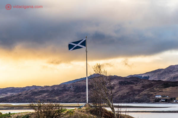 A bandeira escocesa balançando no castelo eilean donan, na Escócia. O céu está alaranjado durante o por do sol, com várias nuvens e montanhas ao fundo.