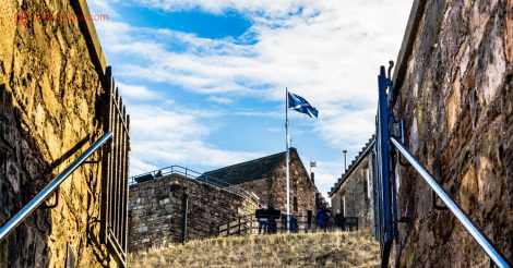 Vista de um ângulo do Castelo de Edimburgo, um dos castelos na Escócia, o mais famoso deles. Fica na cidade de Edimburgo, a mais visitada do país. A foto foi tirada de uma das escadas externas do castelo, de onde é possível ver a bandeira da Escócia balançando no vento, nas cores azul e branca. Ao redor dela, vários outros complexos do castelo. O céu está azul com nuvens brancas.