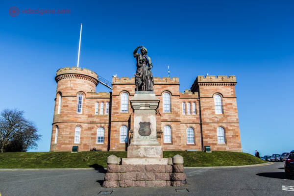 O Castelo de Inverness, nos Highlands da Escócia, com altas vistas da cidade lá fora. O prédio é de uma cor rosada, com torres. Na frente, a estátua de uma mulher. O céu está azul.