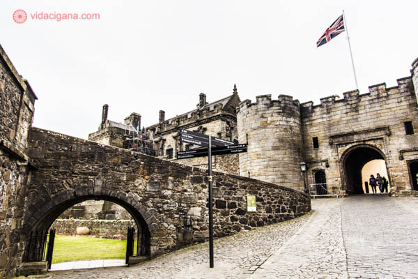 O Castelo de Stirling, na cidade de Stirling, um dos vários castelos na Escócia, cheio de história. Ele fica no alto de uma colina, de onde é possível ver toda a cidade. O castelo é feito de pedras acinzentadas. Na foto é possível ver um ângulo do castelo, logo na entrada, com arcos, pontes e algumas torres. Uma placas explica o caminho por dentro do castelo. O dia está nublado, com o céu branco. Uma bandeira do Reino Unido tremula no alto de um mastro.