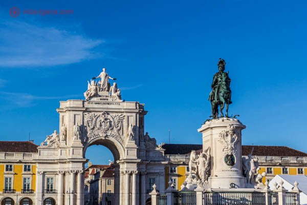 O que visitar em Lisboa: A Praça do Comércio com uma estátua equestre de D. José I em seu meio. Ao fundo, o Arco Triunfal da Rua Augusta. A praça é cercada de prédios amarelos. A foto foi tirada num lindo dia ensolarado.