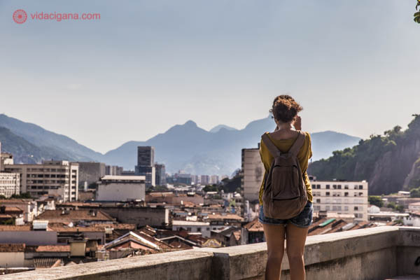 O Morro da Conceição visto do seu alto, com todo o centro do Rio de Janeiro aos seus pés. Uma menina tira foto da vista. O dia está ensolarado, montanhas fecham o horizonte ao fundo.