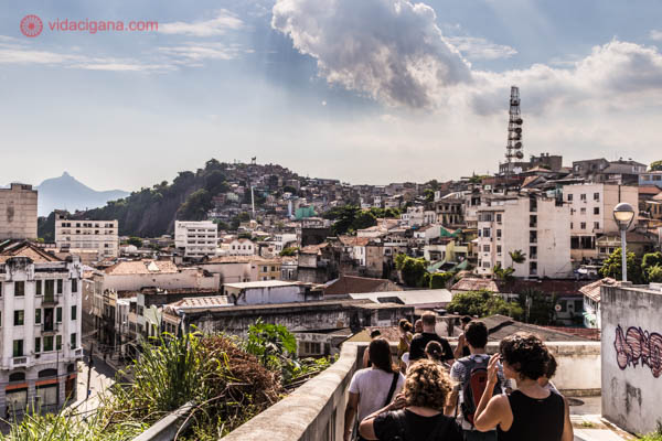 Várias pessoas descem o Morro da Conceição olhando a vista de todo o centro do Rio de Janeiro ao fundo, com suas montanhas, favelas e céu azul.