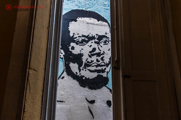 A imagem de Zumbi dos Palmares pintada na parede do Cemitérios dos Pretos Novos, no Rio de Janeiro. A imagem é em preto e branco, em um fundo azul. A foto foi tirada por entre as bandas abertas de uma janela de madeira.