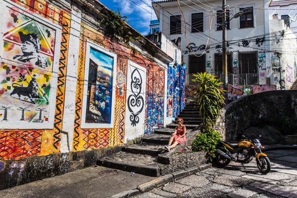 Uma parede repleta de grafites na Pedra do Sal, no Rio de Janeiro. Super coloridos, uma mulher de vestido vermelho se camufla em meio aos murais, sentada na escadaria. Uma moto amarela também decora o lugar.