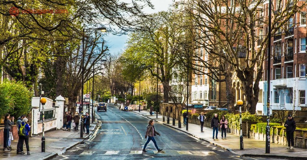 Abbey Road em Londres: Mulher cruza a famosa faixa de pedestres em Abbey Road, na capital inglesa, em um dia de sol e céu azul. Pessoas aguardam sua vez de atravessar a rua. Árvores estão dos dois lados da calçada, com prédios baixos. O asfalto está molhado.