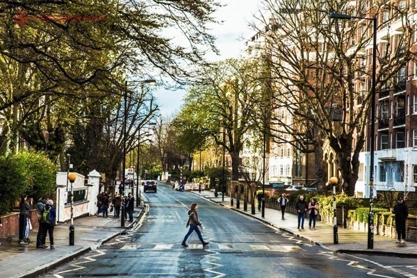 Abbey Road em Londres: Mulher cruza a famosa faixa de pedestres por onde os beatles passaram na foto icônica da capa do álbum chamado Abbey Road. O asfalto está molhado, mas o dia está ensolarado, com árvores verdes nas calçadas, prédios iluminados pelo sol.