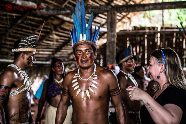 O que fazer em Manaus: Um índio da tribo dessana anda em meio a turistas vestido e pintado de acordo com seus costumes, usando um cocar azul. Ele está dentro de uma oca imensa junto com outros índios. Essa tribo mora perto de Manaus, no estado de Amazonas.