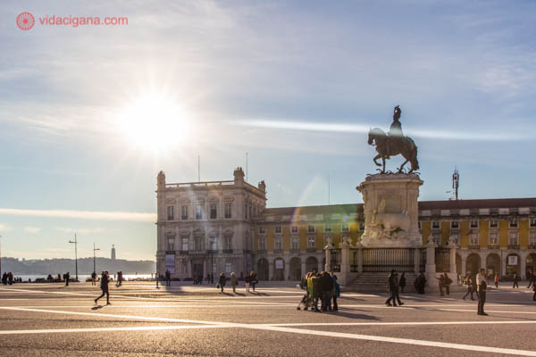 Onde ficar em Lisboa: A Praça do Comércio, em Lisboa, ampla e com algumas pessoas aproveitando os últimos raios de sol do dia. O céu está azul. Uma estátua equestre se encontra no meio da praça, que é cercada por outros prédios baixos. O rio Tejo fica no canto esquerdo da foto.