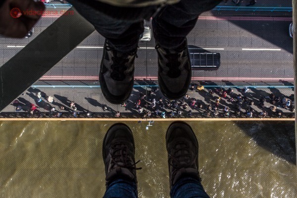 A vista da passarela do topo da mais famosa ponte de Londres, a Tower Bridge, no Rio Tâmisa. Dois pares de pés pisam no vidro com pessoas passando lá embaixo andando na parte pedestre da ponte.