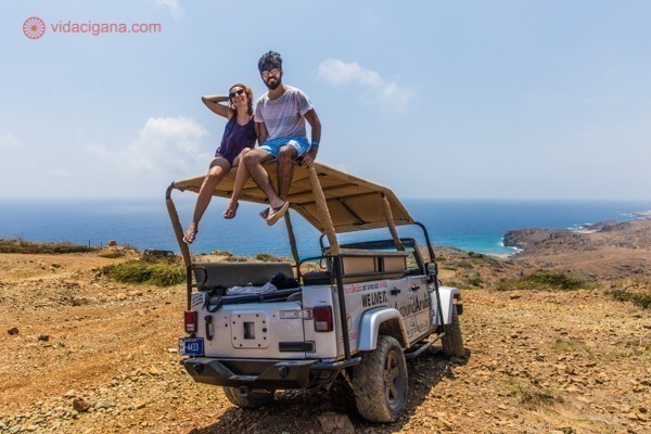 Casal sentado sobre o capô de um veículo 4 por 4 numa paisagem árida com o mar do caribe ao fundo.