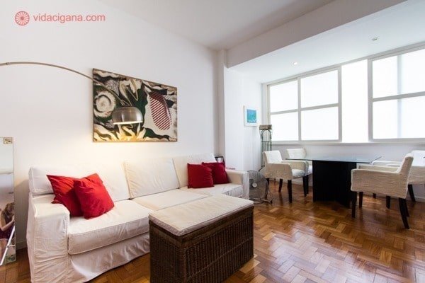 Um sala com sofá branco, almofadas vermelhas, uma luminária caída por cima do sofá, e uma mesa de frente para a janela