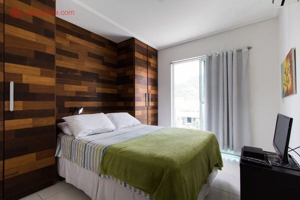 Uma cama com lençóis claros e listrados, cobertor verde claro e com a parede de cabeceira em madeira