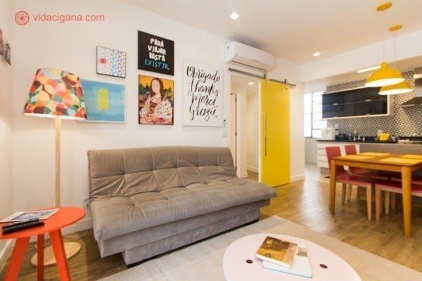 Sala em apartamento em Ipanema, com um sofá cinza, vários quadros acima do sofá, uma cozinha super moderna e colorida, com paredes amarelas