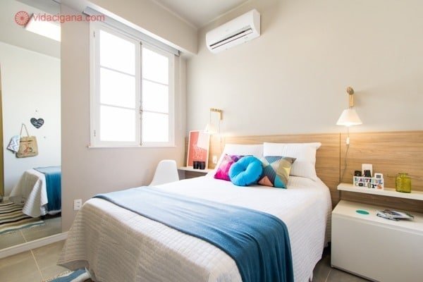 um quarto de airbnb no rio de janeiro com uma cama imensa com lençois brancos e cobertor azul, almofadas coloridas, quarto muito bem iluminado