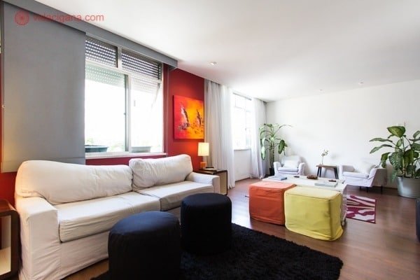 sala de apartamento com sofá claro, almofadas coloridas e algumas azul marinho, paredes brancas e vermelhas, muitas plantas