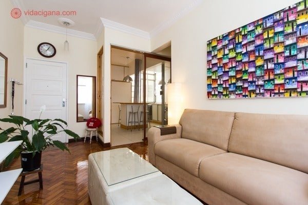 Linda sala de apartamento, com sofá bege, quadro colorido representando as favelas, paredes brancas e cozinha ao fundo com paredes de vidro
