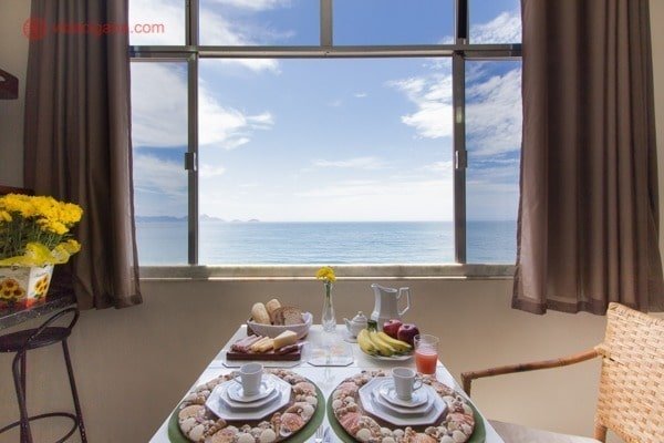 Foto de dentro de um airbnb no rio de janeiro com uma janela aberta de frente para a praia de copacabana, uma mesa montada na frente da janela com café da manhã