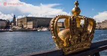 O que fazer em Estocolmo: A Ponte Skeppsholm em primeiro plano, com sua estrutura metálica e uma coroa dourada em seu gradil, com o Palácio de Estocolmo em seu fundo. O céu está azul, com nuvens brancas. Entre a ponte e o palácio fica o mar.