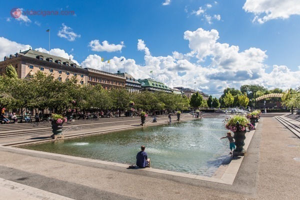O que fazer em Estocolmo: O Jardim do Rei, com sua enorme fonte, parecendo uma piscina, com várias pessoas sentadas em suas margens. O quarteirão é cercado por árvores e prédios coloridos.