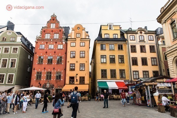 O que fazer em Estocolmo: A Praça Principal de Gamla Stan, em Estocolmo, com seus prédios baixos e coloridos, em tons terrosos que vão do laranja ao amarelo. Muitas pessoas estão paradas em meio à praça