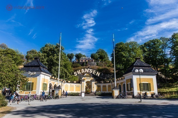 O que fazer em Estocolmo: O Skansen, um dos primeiros museus a céu aberto no mundo, com sua entrada amarela, com o letreiro escrito Skansen, e lá ao fundo, árvores verdes e céu azul.