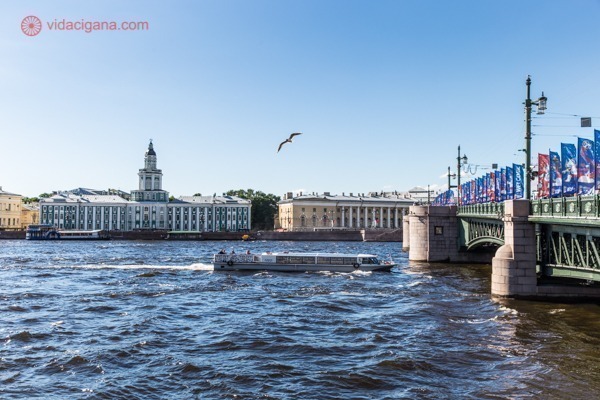 Onde ficar em São Petersburgo: O rio Neva, em São Petersburgo, de tonalidade azul escuro, com uma ponte por onde um barco comprido atravessa. Do outro lado do rio estão vários prédios históricos, de várias cores. O céu está azul e as gaivotas estão voando.