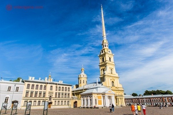 Onde ficar em São Petersburgo: O interior da Fortaleza de São Pedro e São Paulo, com sua igreja dourada e com uma torre alta e fina. Vários prédios ao redor amarelos. O céu está azul com poucas nuvens brancas.