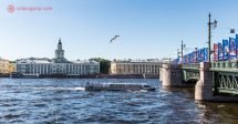 O que fazer em São Petersburgo: A margem do Rio Neva, com um barco passando por baixo da Ponte do Palácio, repleta de bandeiras da Copa do Mundo 2018. Do outro lado da margem, o museu Kunstkamera, com suas paredes verdes. O céu está azul e uma gaivota voa no céu.