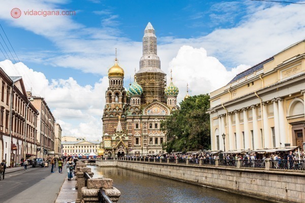 O que fazer em São Petersburgo: A Catedral do Sangue Derramado na beira de um dos canais de São Petersburgo, com suas paredes coloridas e domos dourados. O céu está azul com nuvens brancas.