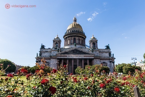 O que fazer em São Petersburgo: A Catedral de Santo Isaac, com a Praça de Santo Isaac em sua frente, com um jardim cheio de rosas vermelhas. A igreja é imensa e possui uma cúpula banhada a ouro. O céu está azul.
