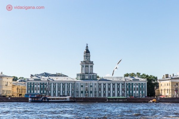 O que fazer em São Petersburgo: O Kuntskamera, museu na beira do Rio Neva construído por Pedro, o Grande. Sua paredes verdes e colunas brancas se destacam na arquitetura a beira rio. O dia está ensolarado e uma gaivota voa na frente do museu.
